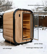 Sauna extérieur en bois avec porte vitrée ouverte, cadre noir, éclairage LED intérieur et sol enneigé